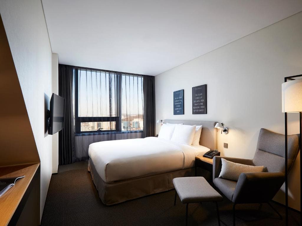 Modern hotel room with a large bed, two wall lamps, an armchair with an ottoman, a floor lamp, a wall-mounted TV, and a window with sheer curtains letting in natural light.