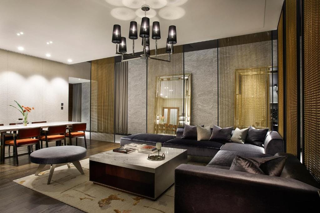 Modern living room with a large sectional sofa, low coffee table, and ottoman on a patterned rug. Dining area with red chairs is visible, and decorative gold screens, mirrors, and a black chandelier enhance the space.