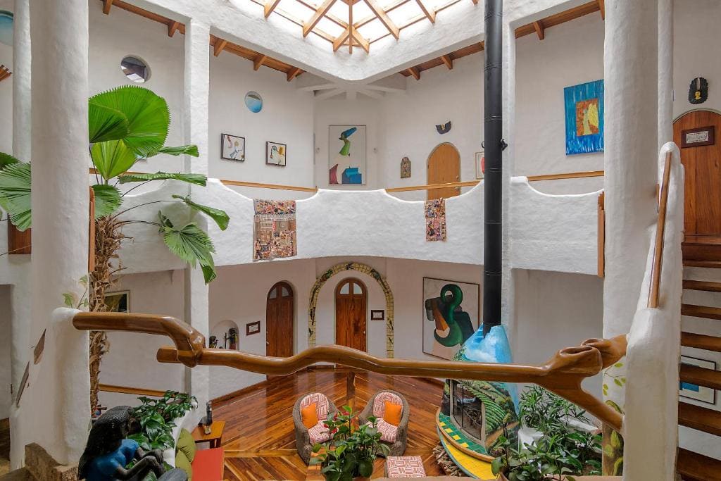 A bright, spacious interior with white curved walls, wooden floors, skylight ceiling, indoor plants, colorful artwork, and a cozy seating area with two orange chairs, surrounded by natural decor and unique architectural details.