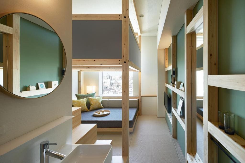 A modern, compact room features a loft bed with wooden frames, a cozy seating area by a large window, pastel green walls, a round mirror above a sink, and built-in shelves along the passageway.