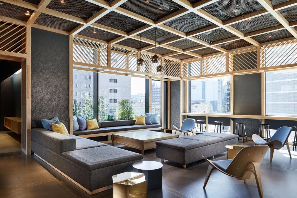 Modern lounge with gray sectional sofas, wooden accents, and large windows letting in natural light. Decorative pendant lights hang from a geometric ceiling. City buildings are visible outside.