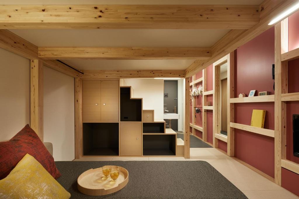 A cozy room with light wood beams, built-in storage under stairs, a small round table with two glasses, gray and red cushions, and red accent walls, creating a modern Japanese-inspired interior.