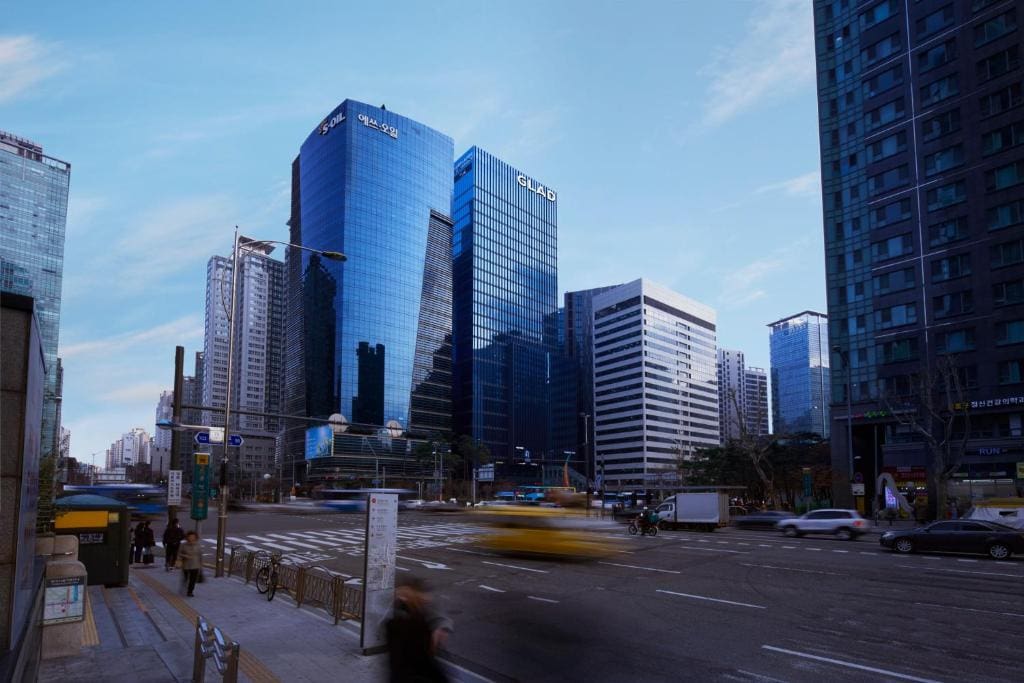 A city street with blurred cars and people in motion, surrounded by modern glass skyscrapers under a clear blue sky. Bicycles are parked along the sidewalk.