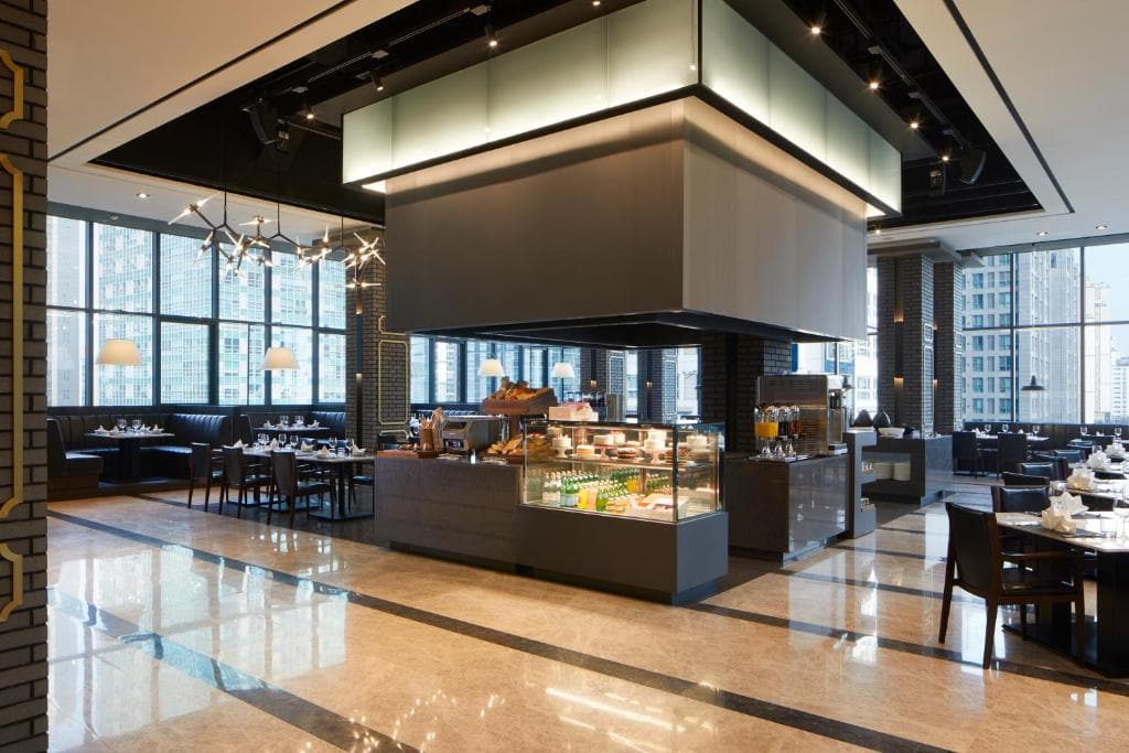 A modern, spacious restaurant with large windows, dark furniture, and a central buffet counter displaying food items. The room features marble floors, black accents, and city buildings visible through the windows.