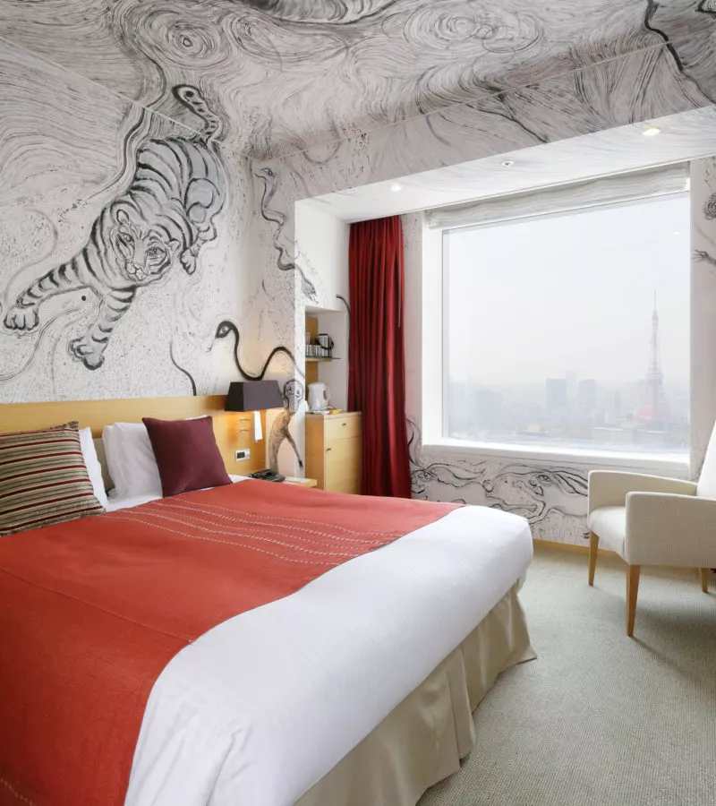 A hotel room with a double bed, red and white bedding, a large window with a city view, and detailed wall art featuring a tiger and swirling patterns. There is a beige armchair and wooden furniture by the window.