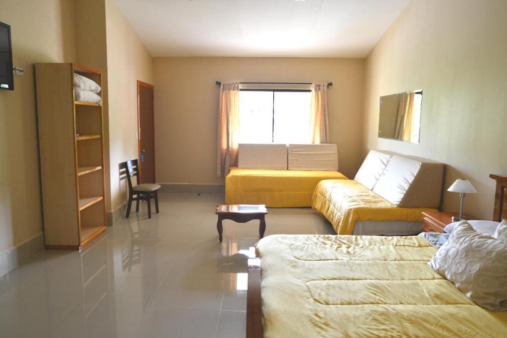 Bright, spacious bedroom with yellow bedding, a double bed, sofa, and cushioned bench by the window. Room includes a wall-mounted TV, open shelving, chair, coffee table, and large mirror. Cream walls and glossy floor tiles.