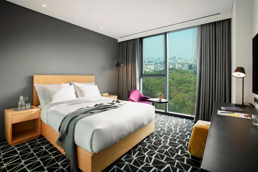 A modern hotel room with a large bed, nightstands, desk, and chair. Floor-to-ceiling windows reveal a city skyline and green trees outside. The decor is minimalist, with geometric carpet and neutral colors.