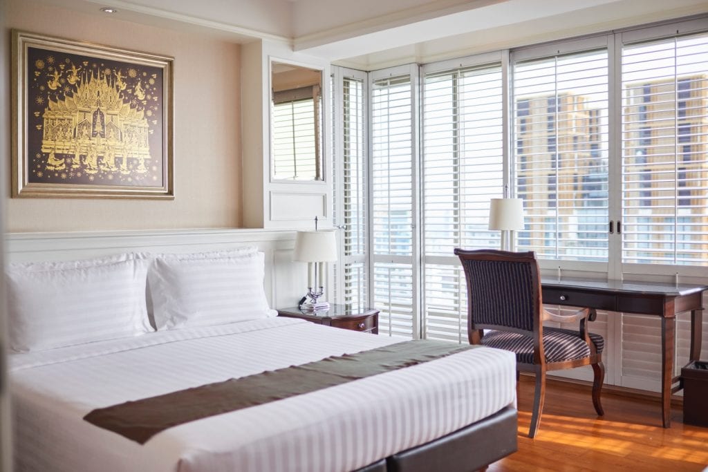 A bright hotel room with a neatly made bed, bedside tables with lamps, a wooden desk and chair by large windows with white blinds, and a gold-framed artwork on the wall.