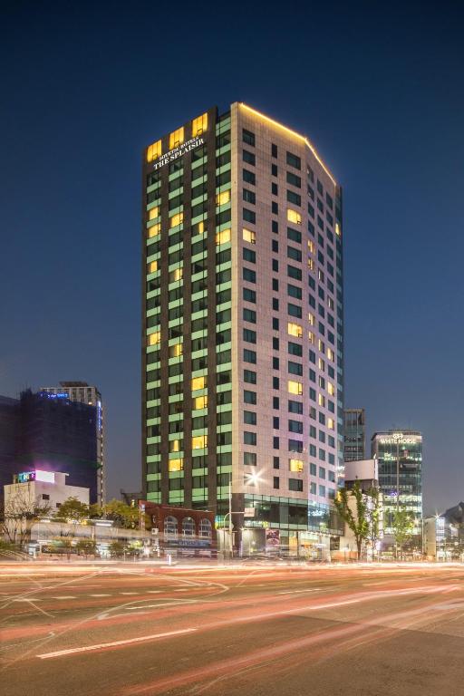 A tall, modern hotel building with illuminated windows and lights outlining the roof, stands against a clear night sky, with blurred car lights on the street in front.