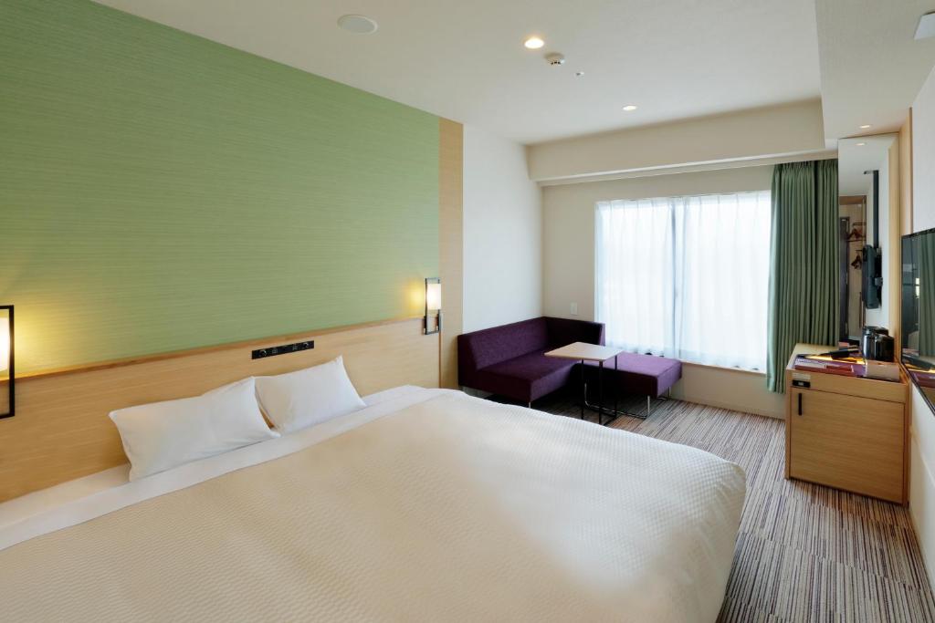Modern hotel room with a king-size bed, green accent wall, wall lamps, a purple sofa by the window, desk with a TV, and striped carpet flooring. Natural light enters through a large window with light curtains.
