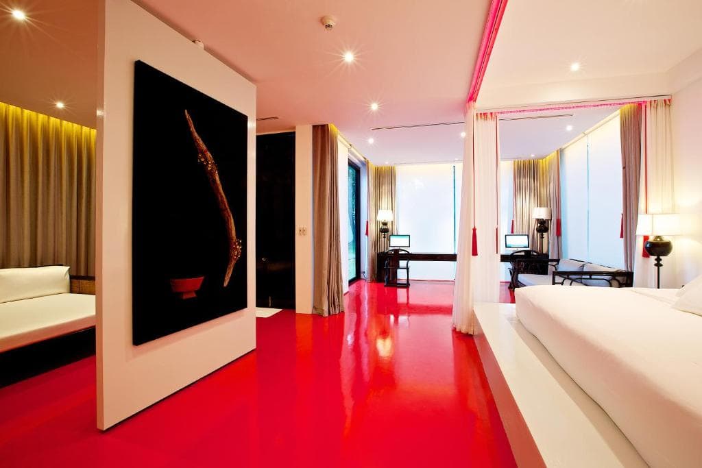 A modern hotel room with glossy red floors, white walls, a large bed with white linens, sheer curtains, contemporary furniture, dramatic lighting, and a large abstract artwork on a partition wall.