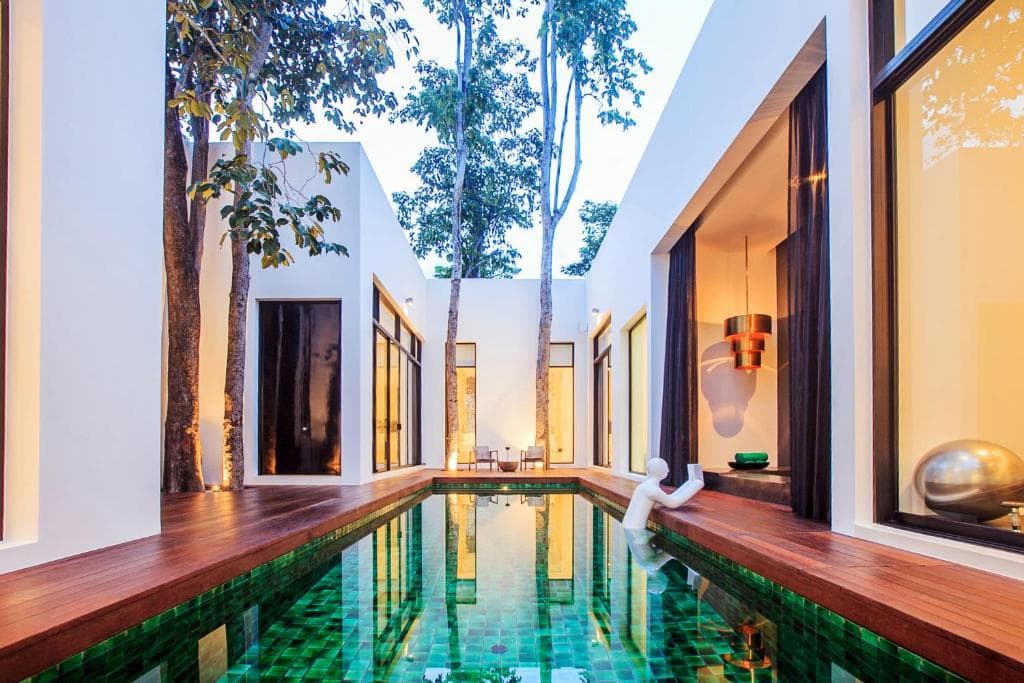 Modern courtyard with a rectangular green-tiled pool surrounded by white buildings, large windows, and tall trees growing through the wooden deck. A white abstract sculpture sits at the pool’s edge. Warm indoor lighting is visible.