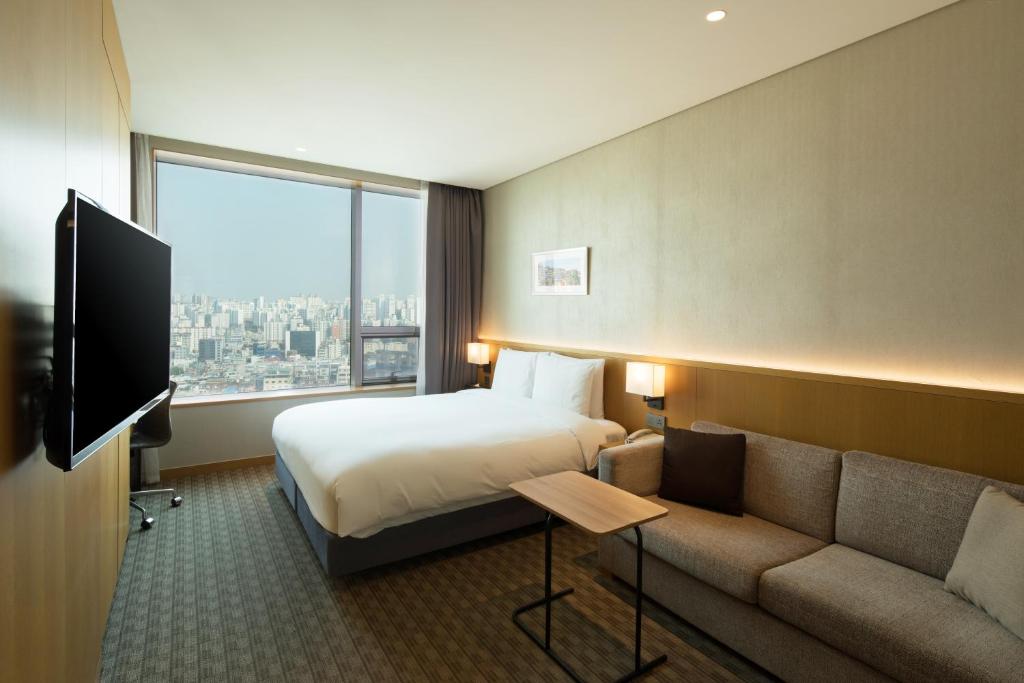 A modern hotel room with a large bed, a gray sofa, a small wooden table, wall-mounted TV, and a floor-to-ceiling window offering a city view. The room is well-lit with neutral-toned decor.