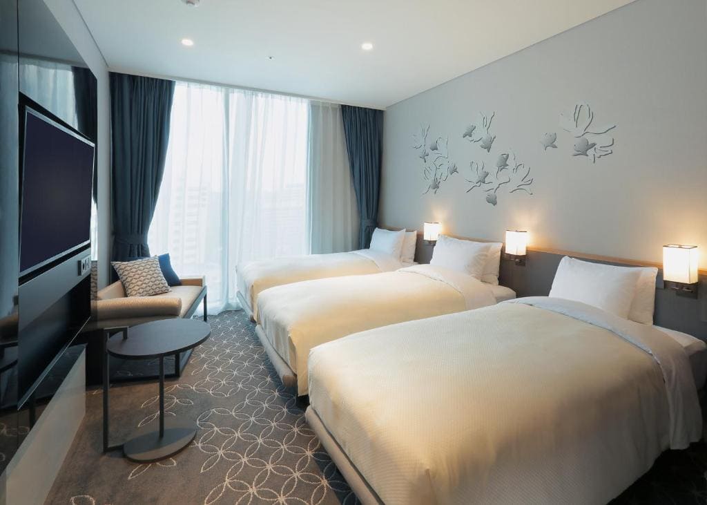 A modern hotel room with three single beds, white bedding, wall-mounted lamps, a flat-screen TV, a small table with chairs, floor-to-ceiling windows with sheer curtains, and decorative wall art above the beds.