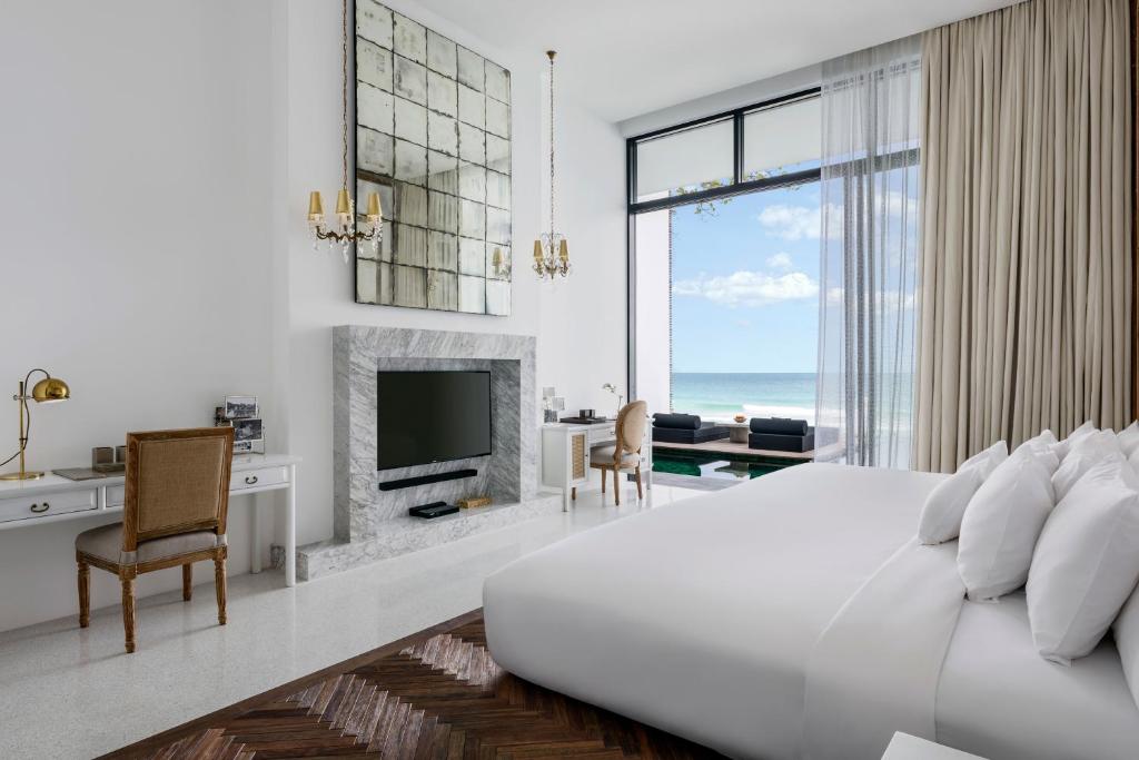 Modern, bright hotel room with a large bed, marble fireplace, TV, elegant desk, armchair, and tall windows with sheer curtains. Ocean view visible through the windows, creating a serene and airy atmosphere.