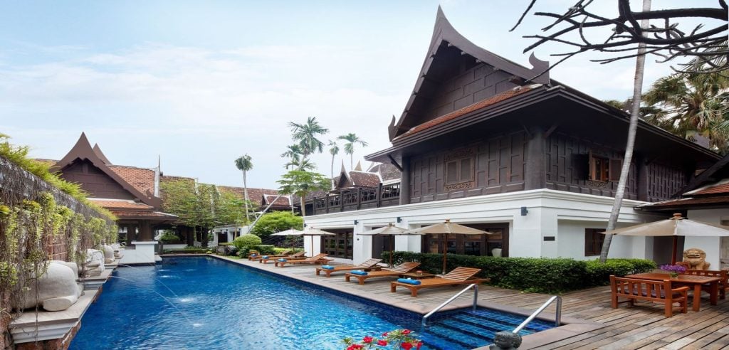 A luxurious villa with traditional Thai architecture, a private blue-tiled swimming pool, wooden deck with lounge chairs, and white umbrellas, surrounded by lush greenery and statues.