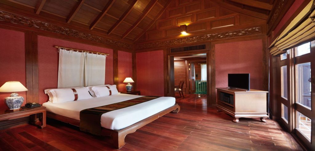 A cozy wooden bedroom with a large bed, bedside tables with lamps, a flat-screen TV on a wooden cabinet, and a window letting in natural light. The room features warm lighting and wood-paneled walls and floor.