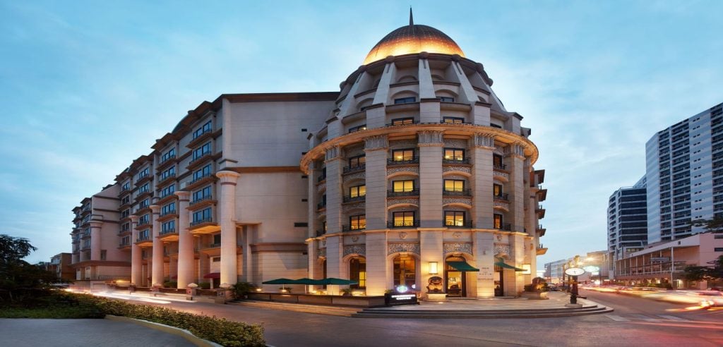 A grand, multi-story hotel building with ornate columns and a domed corner tower is illuminated at dusk, with streaks of car lights on the street and modern skyscrapers in the background.