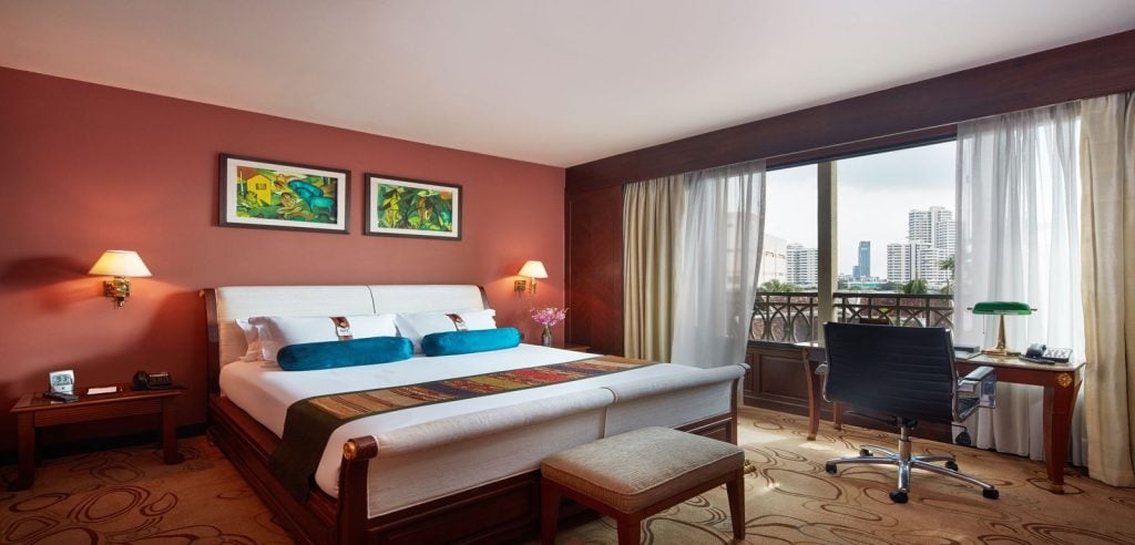 A hotel room with a large bed, colorful pillows, artwork on a red accent wall, bedside lamps, a desk with a chair, and a window with city buildings visible outside.