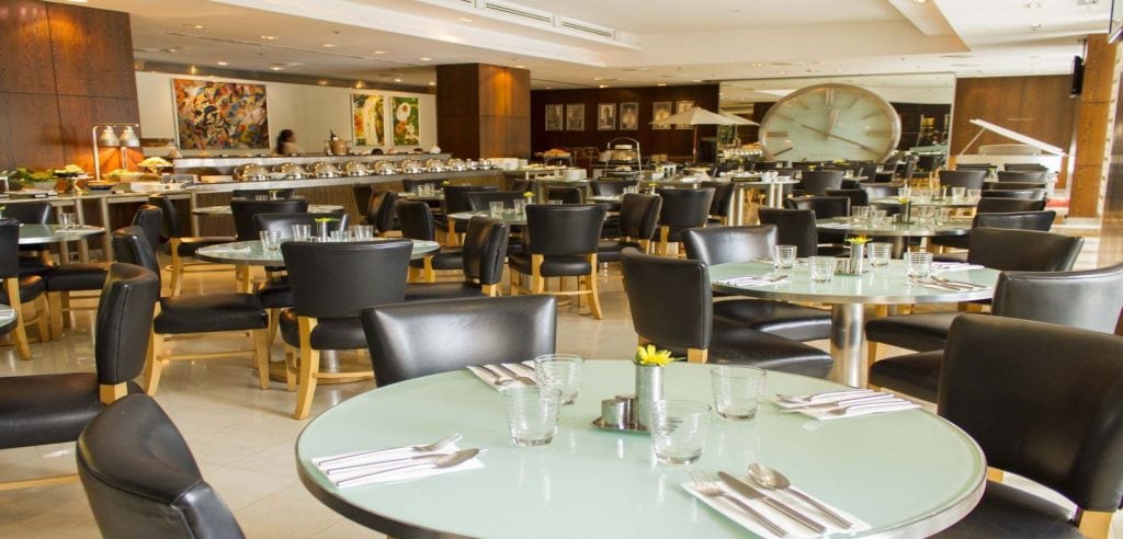 A modern restaurant dining area with round tables set for guests, black chairs, and glass tabletops. The background shows a buffet area and colorful artwork on the walls. The restaurant appears empty.