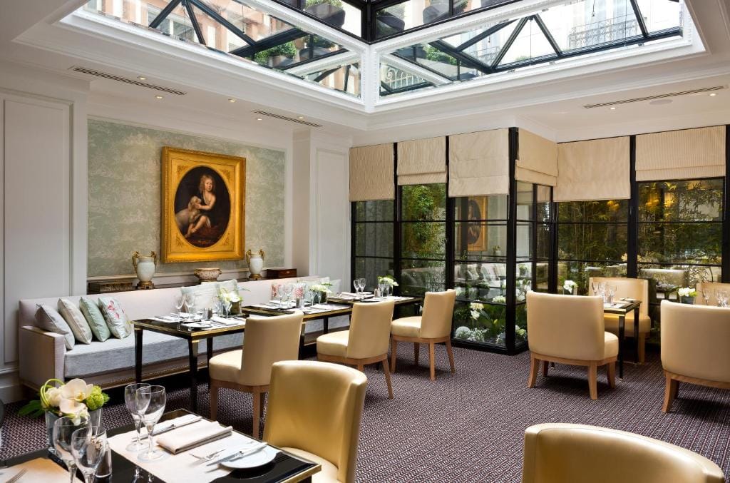 Elegant restaurant with cream chairs, set tables, and glass ceiling. Large windows offer garden views. A classical portrait hangs on the wall above a cushioned bench with pillows. Bright, airy, and sophisticated atmosphere.