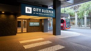 Entrance to the City Sleeper at the Royal National Hotel, with illuminated signs, a glass door, and a partial view of a red double-decker bus and trees on the street outside.