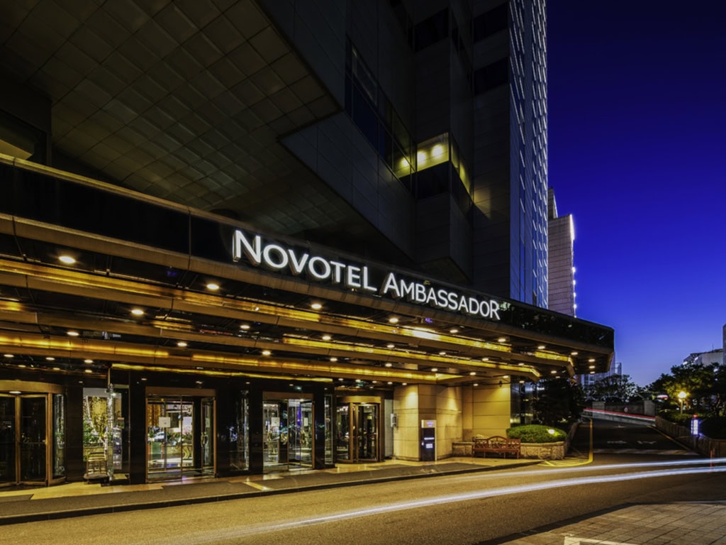Exterior view of the Novotel Ambassador hotel at night, with its brightly lit entrance and signage, modern glass doors, and a dark blue evening sky in the background.