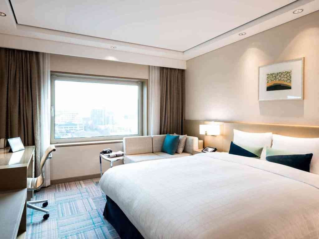 A modern hotel room with a large bed, white bedding, a sofa by the window, a desk with a chair, and city buildings visible outside. The room has neutral colors and minimalistic decor.