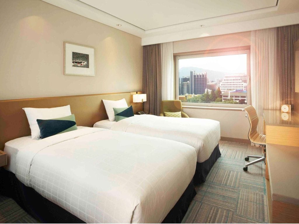 A modern hotel room with two neatly made twin beds, a desk and chair by the window, a lamp, wall art, and a large window offering a view of city buildings and greenery outside.