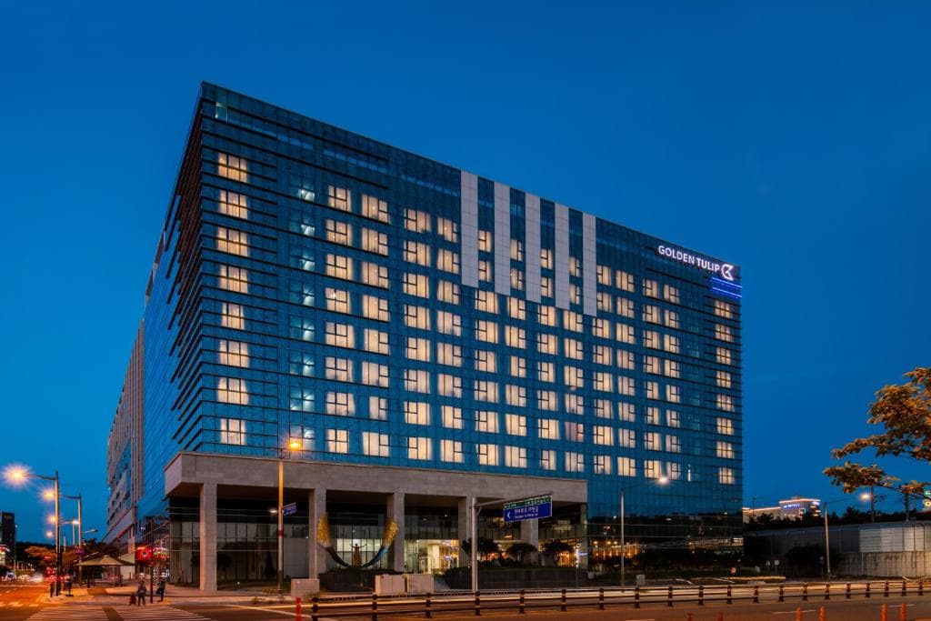 A modern, multi-story hotel building with many windows lit up at dusk. The hotel has a glass facade and a sign that reads Golden Tulip. Streetlights and a few cars are visible in the foreground.