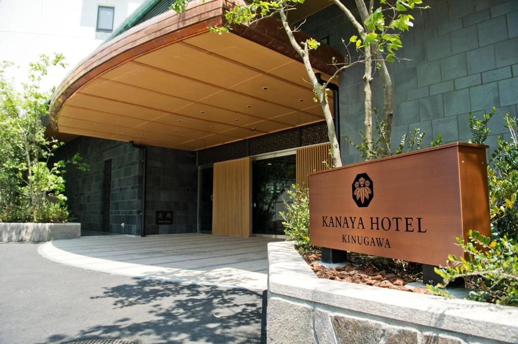 The entrance of Kanaya Hotel Kinugawa features a modern wooden canopy, stone walls, a sliding door, and a brown sign with the hotel’s name and logo surrounded by green plants.