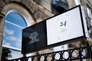 A rectangular sign mounted on a black metal fence displays HOTEL GALLERY BAR GARDENS on a black background and 24 ROYAL TERRACE on a white background, with a stone building in the background.