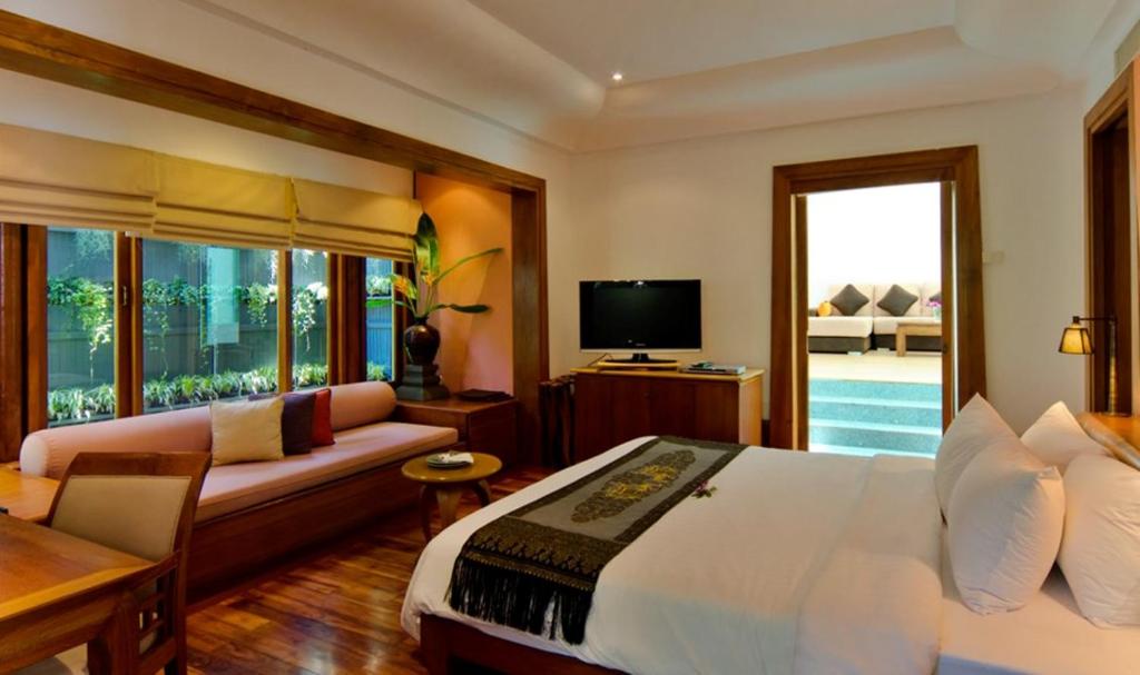 A cozy hotel room with a large bed, a cushioned seating area by the window, a flat-screen TV, wooden furniture, and a view of a private pool and outdoor seating through a glass door.