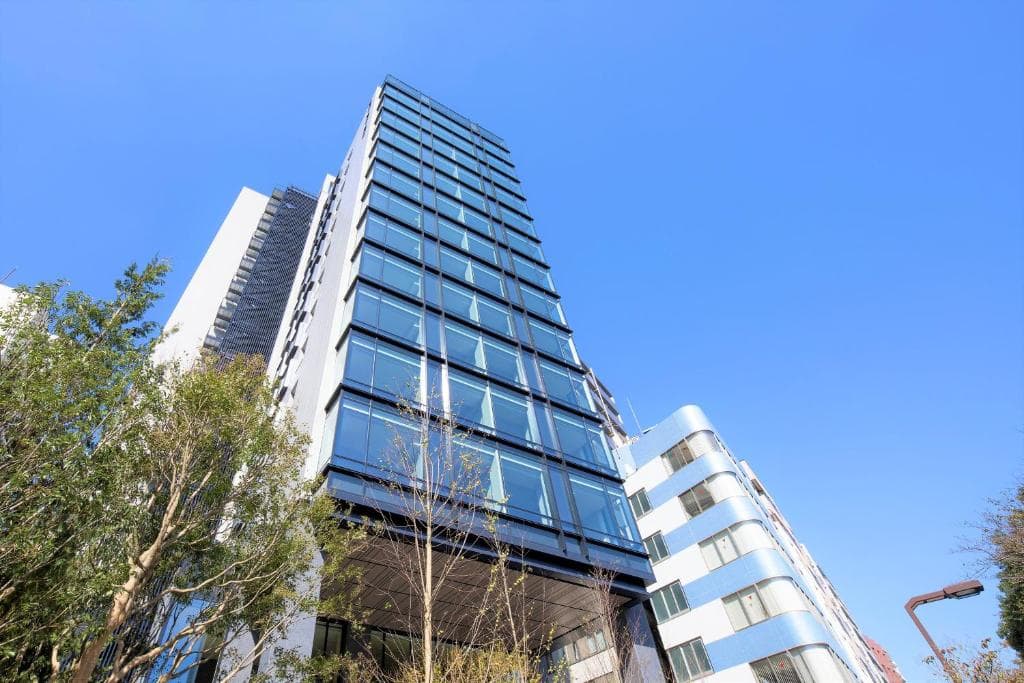 A modern glass office building with large windows rises against a clear blue sky, with trees and neighboring buildings visible at the sides.