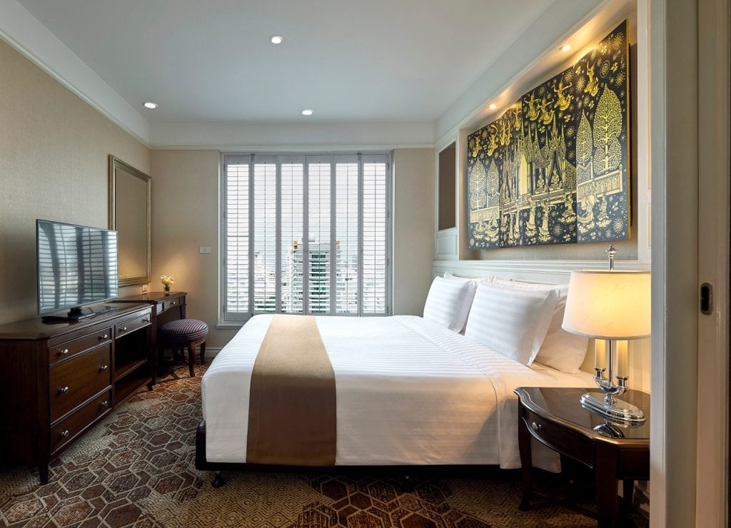 A modern hotel room with a large bed, white linens, brown runner, bedside tables with lamps, a TV on a wooden dresser, a desk with a chair, a large window with blinds, and a gold-accented artwork above the bed.