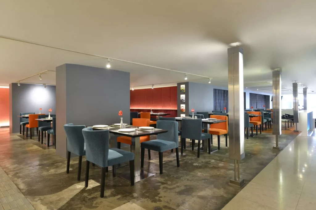 A modern restaurant interior with neatly set tables, blue and orange upholstered chairs, gray walls, and contemporary lighting. The atmosphere is clean and spacious with minimalist decor.