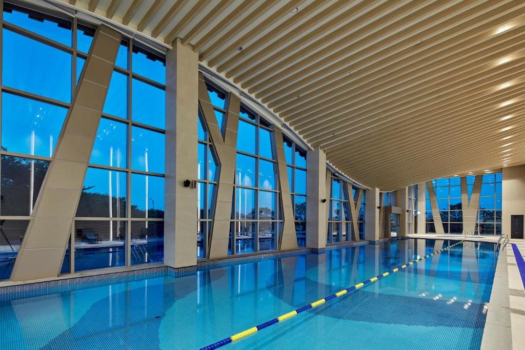 Indoor swimming pool with clear blue water, lane dividers, and large floor-to-ceiling windows. The ceiling features curved beams, and the view outside shows trees and a blue sky at dusk.