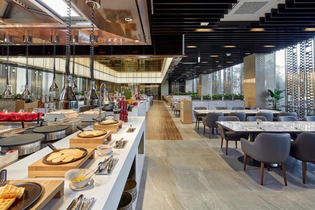 A modern, spacious buffet restaurant with neatly arranged food on counters to the left and elegant dining tables with gray chairs to the right. Large windows let in natural light, and green plants decorate the space.
