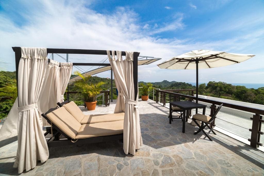 A rooftop terrace with a lounge bed under a canopy, potted plants, a table with two chairs, and a large umbrella. The area overlooks greenery and has a view of the distant ocean under a partly cloudy sky.
