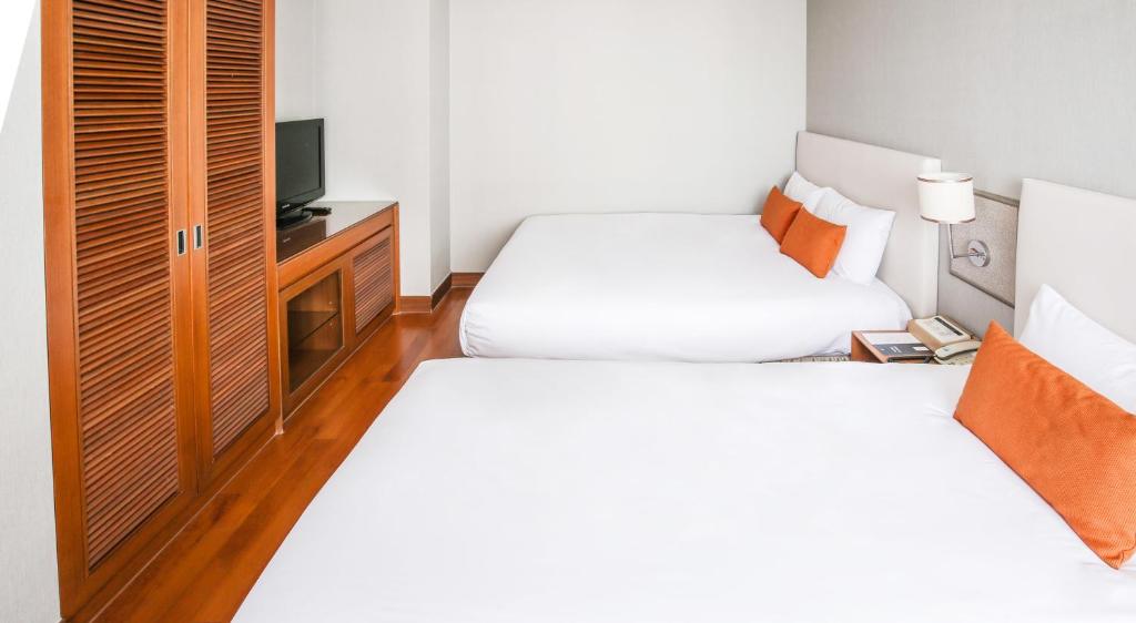 A hotel room with two large beds, each with white bedding and orange pillows, a wooden floor, a wooden wardrobe, a TV on a cabinet, and a nightstand with a phone and lamp. The walls are light-colored.