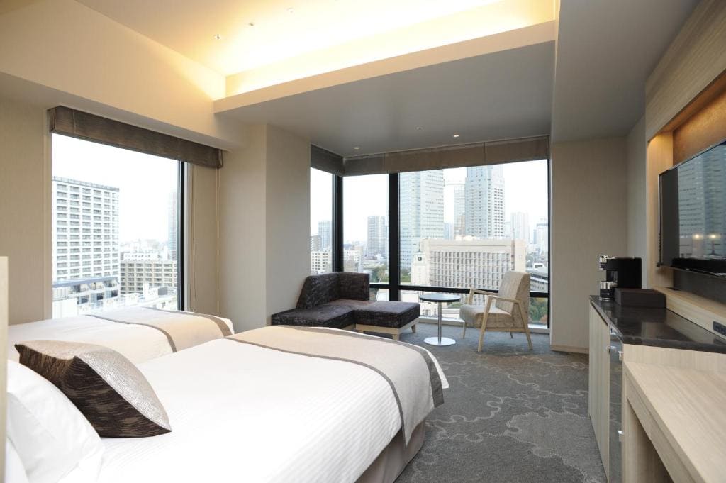 A modern hotel room with two beds, a sofa, a chair, floor-to-ceiling windows offering a city view, and a wall-mounted TV. The room has neutral decor and ample natural light.