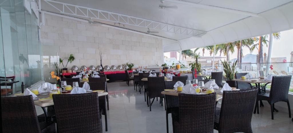 Outdoor restaurant terrace with wicker chairs and set dining tables, white cloth napkins, and glassware. Buffet tables with food are at the back. Large windows and palm trees overlook a seaside view.