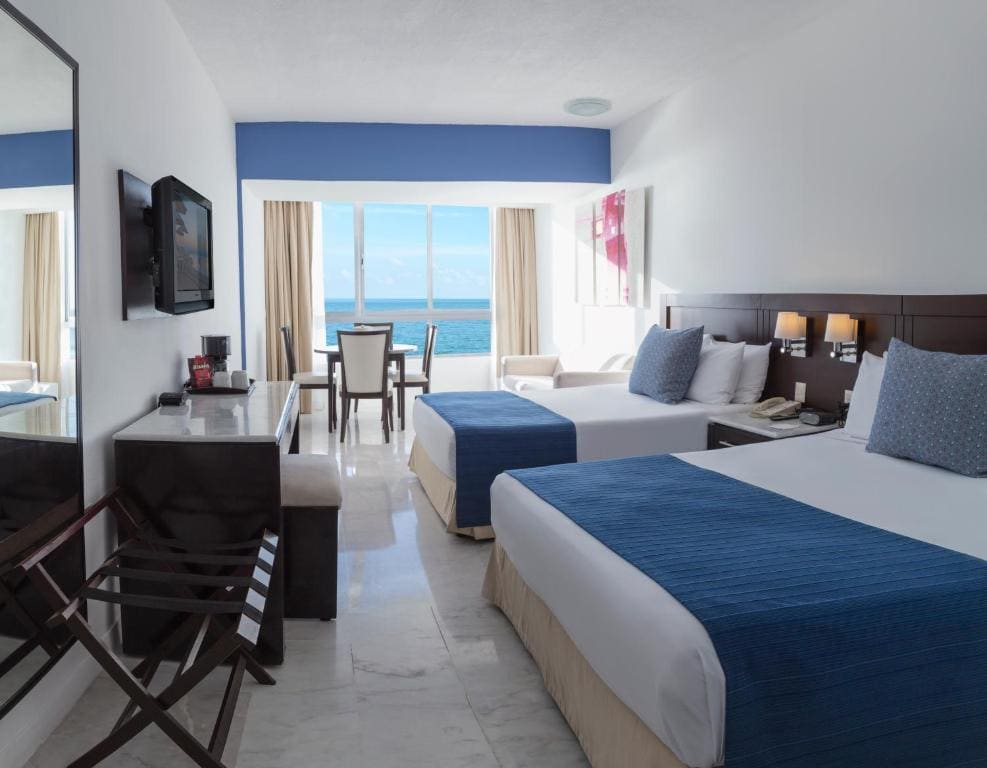 A bright hotel room with two double beds, blue and white bedding, a desk with a TV, and a large window overlooking the ocean. There is a small dining area near the window and marble flooring throughout the room.