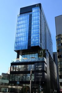 A modern glass-fronted office building with several stories, prominent signage, and reflective blue windows, standing against a clear blue sky on a sunny day.