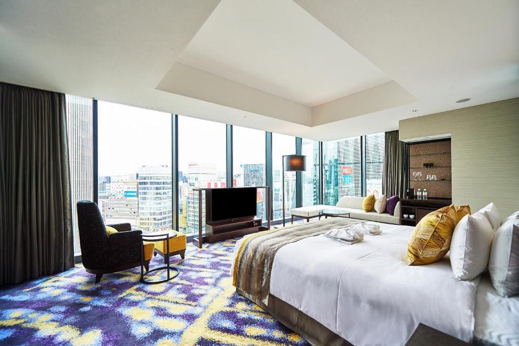 A modern hotel room with a large bed, gold and white pillows, a TV, armchair, table, and a sofa by floor-to-ceiling windows overlooking a city skyline. The room has a colorful patterned carpet.