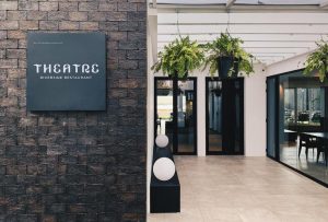 Entrance to THEATRE Riverside Restaurant with a modern sign on a brick wall, potted plants hanging above, and glass doors leading to a bright, contemporary interior with tables and chairs visible inside.