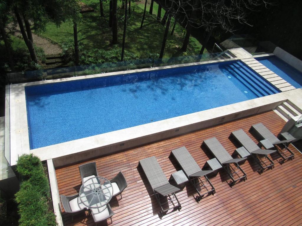 A rectangular outdoor swimming pool next to a wooden deck with several lounge chairs and a round glass table with chairs, surrounded by greenery and trees.