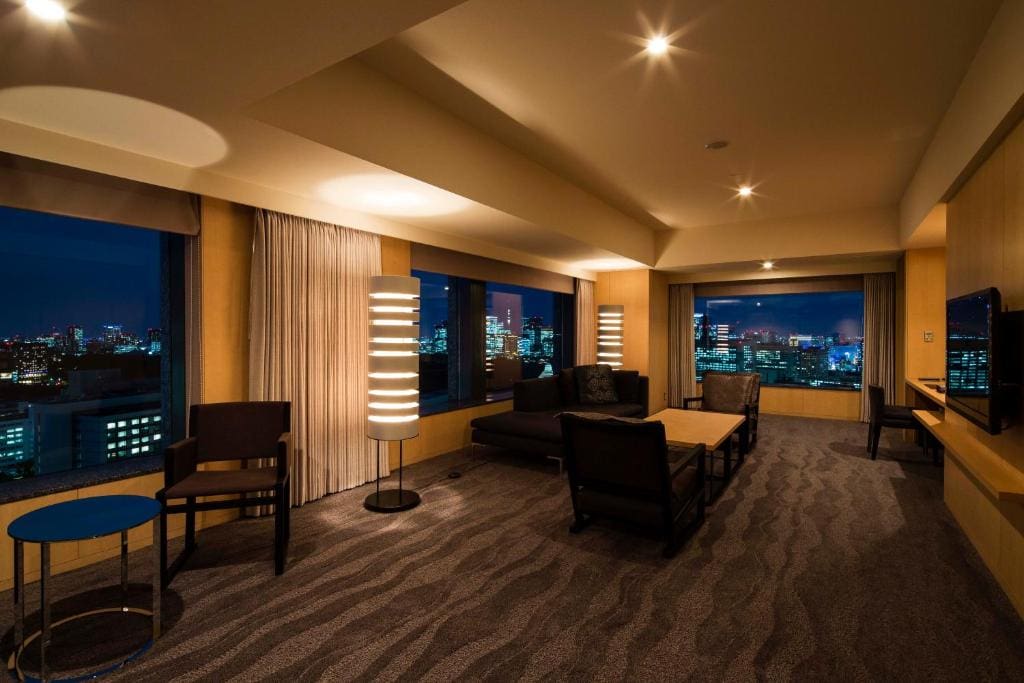Spacious modern hotel suite at night with large windows showing a city skyline, contemporary furniture, two tall floor lamps, a wall-mounted TV, and patterned carpet. The room is warmly lit and elegantly decorated.