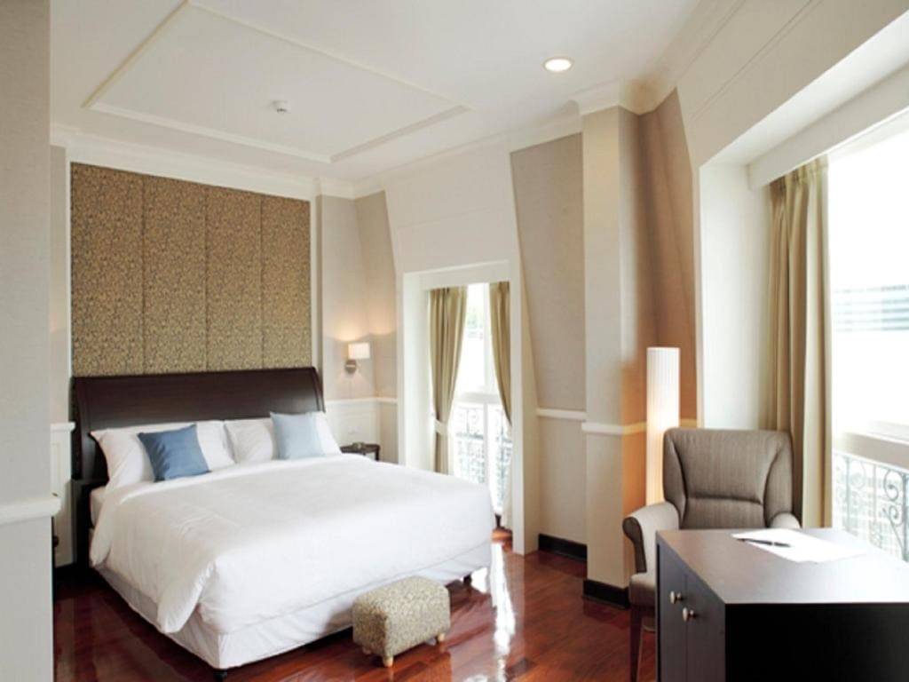 A bright, modern bedroom with a large bed, white bedding, blue and gray pillows, an upholstered chair, a small desk, wood floors, tall windows with curtains, and soft lighting.