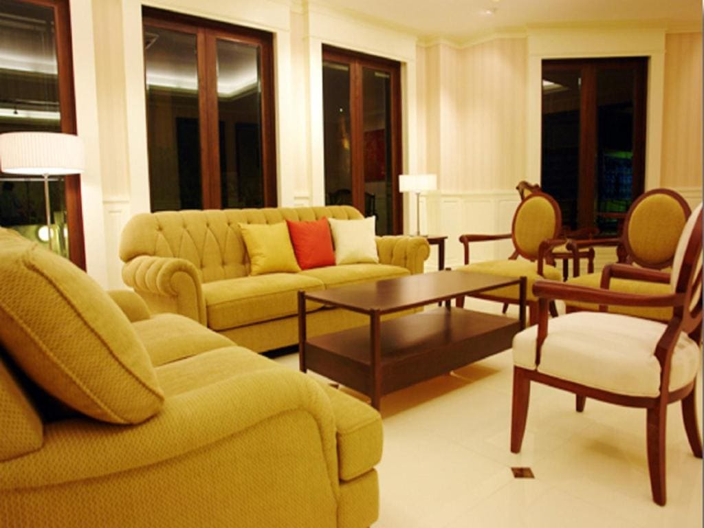 A stylish living room with beige sofas, two wooden armchairs with cream cushions, a brown coffee table, and a floor lamp, all set against large windows and cream-colored walls.